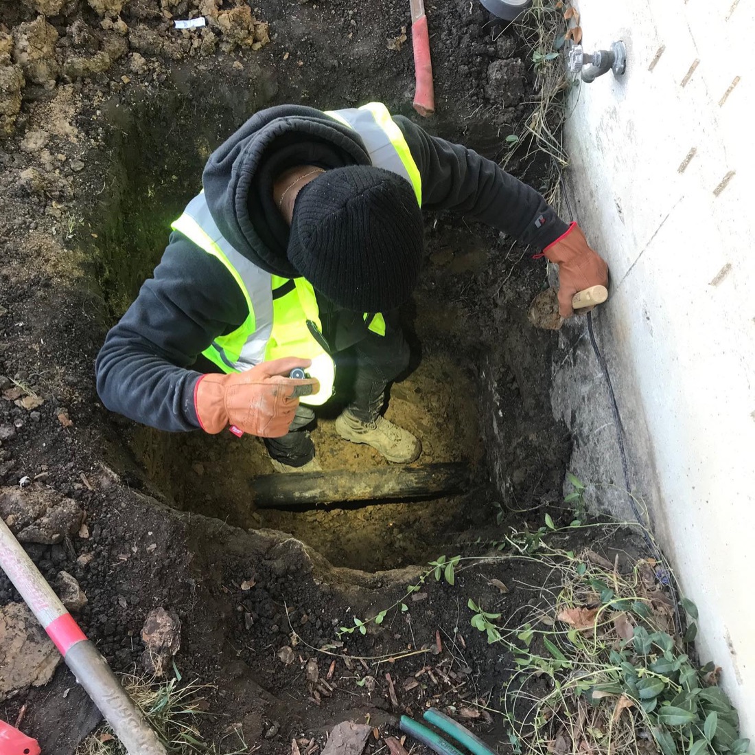 Technician in a high-visibility vest working in a hand-dug hole beside a foundation